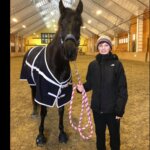 Horse with Cavallo hoof boots, wearing a rug, standing in an indoor arena with person holding reins.