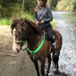 Rider on a horse wearing green Cavallo hoof boots by a scenic riverside, showcasing durable equine gear.