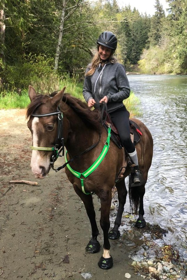 Rider on a horse wearing green Cavallo hoof boots by a scenic riverside, showcasing durable equine gear.