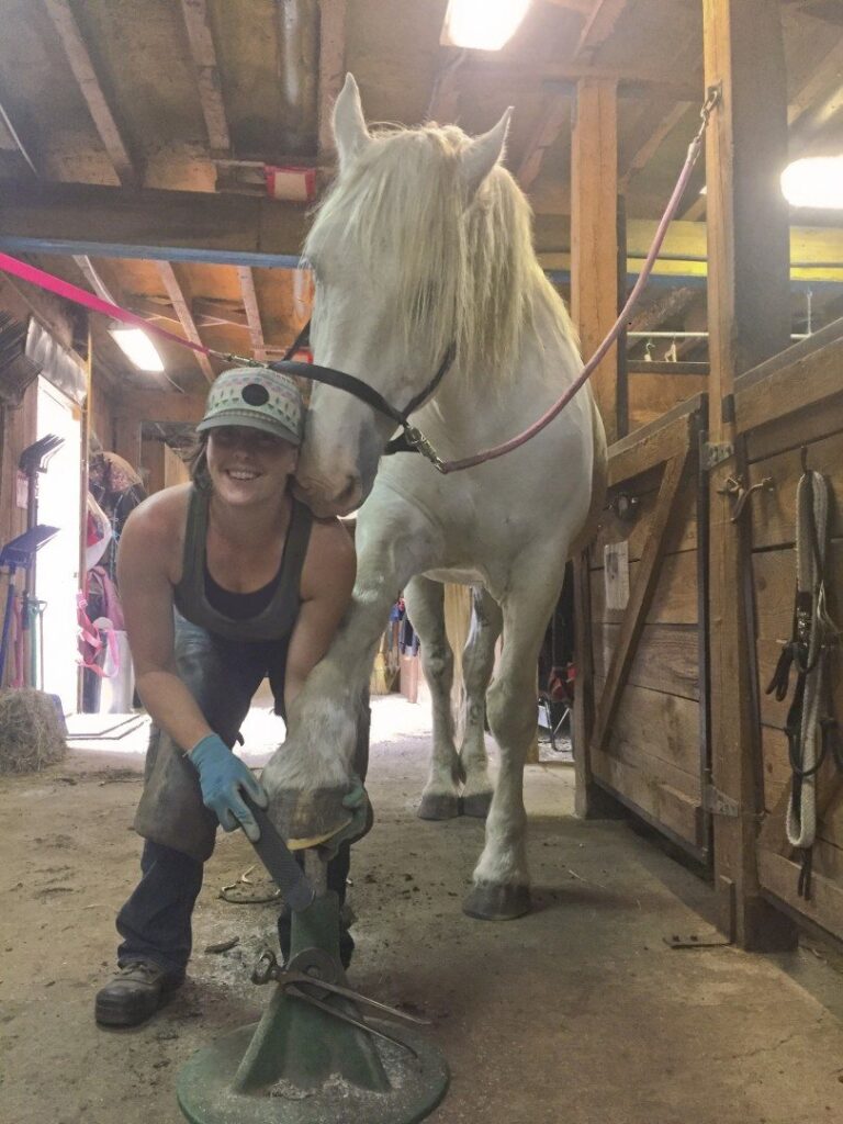 A trimmer working on a hoof in a barn aisle, a white horse beside her